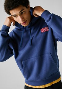 Sudadera azul marino hecha de un tejido suave, con un bolsillo frontal, cordones y un parche con el logo rojo de "Red Bull Racing" en el pecho.