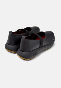 Black leather shoes with a rounded shape, elastic strap, and a rubber sole with textured yellow grips. Interior features a red lining.