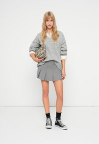 Szary, za duży sweter, szara plisowana spódnica mini, czarne skarpetki i trampki za kostkę. Trzyma torebkę w wężowy wzór. Neutralne tło.