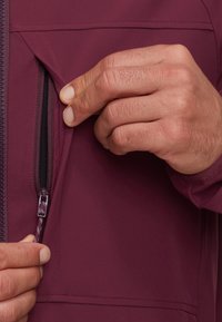 Des mains ouvrant une petite poche verticale sur une veste bordeaux, montrant les détails de la fermeture éclair et du tissu.