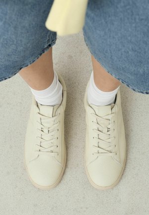 Benen in blauwe spijkerbroeken met witte sokken en lichtbeige vetersneakers op een lichtgrijze vloer.