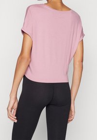 Růžové krátké tričko vyrobené z měkké látky s ohrnutými krátkými rukávy a kulatým výstřihem, v kombinaci s černými legínami.