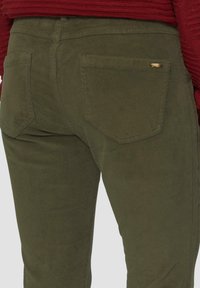 Olivgrüne Cordhosen aus strukturiertem Stoff, mit Fünf-Taschen-Design, sichtbaren Nähten und einem kleinen Metallelement auf der Gesäßtasche.