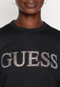 Gros plan sur une personne portant un sweat-shirt noir avec "GUESS" écrit en grandes pierres précieuses brillantes sur la poitrine, montrant uniquement les lèvres et le cou.