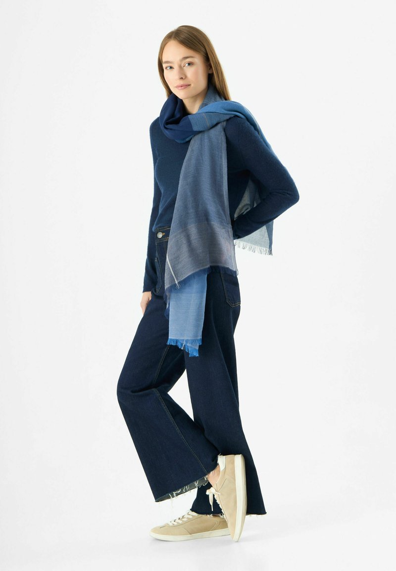 Pull en laine bleu marine, écharpe bleu clair avec des bords effilochés, jeans larges noirs et baskets beiges. Présente des nuances de bleu variées.