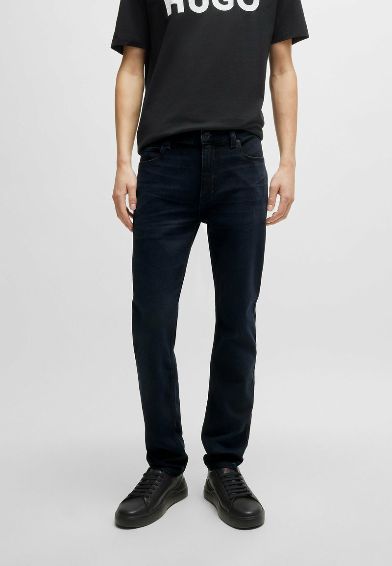 Jeans de mezclilla azul oscuro con un corte recto, que cuentan con cinco bolsillos y detalles de costura sutiles, combinados con zapatillas negras y una camiseta negra.