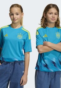 Två barn bär blåa Spanien landslagströjor i fotboll med gula accenter och grafik. Tröjorna har korta ärmar och en avslappnad passform.