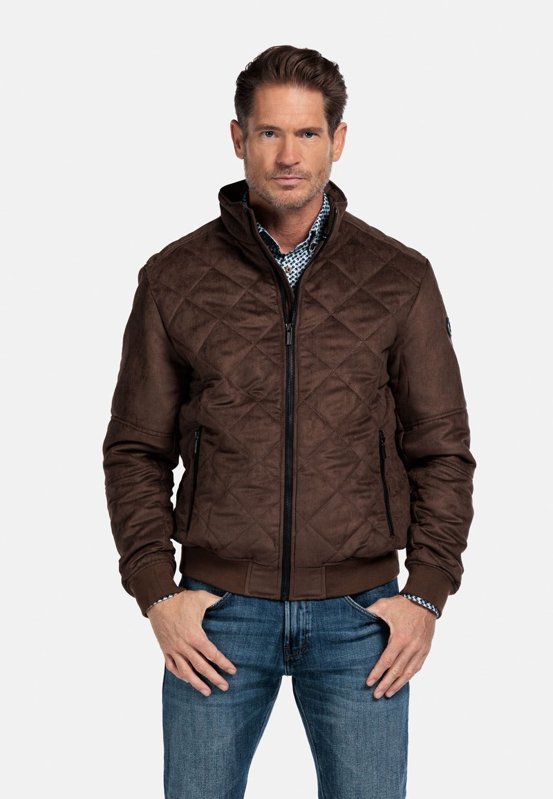 Bruine gewatteerde bomberjack met een hoge kraag, een rits aan de voorkant en zijzakken. Combinatie met blauwe jeans en een bedrukte shirt eronder.