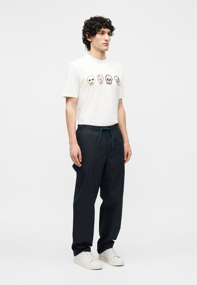 Νεαρός άντρας στέκεται φορώντας λευκό μπλουζάκι με τέσσερις πολύχρωμες απεικονίσεις κρανίων, σκούρο παντελόνι με κορδόνι και λευκά αθλητικά παπούτσια σε απλό φόντο.