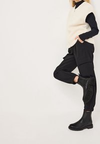 Chaleco de piel sintética crema sobre un cuello de tortuga negro de canalé, combinado con pantalones cargo negros y botines negros con suelas texturizadas.