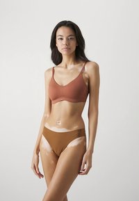 Brun sømløs bralette og matchende bikinibottom på en modell. Stoffet ser glatt ut med en matt finish. Justerbare BH-stropper er synlige.
