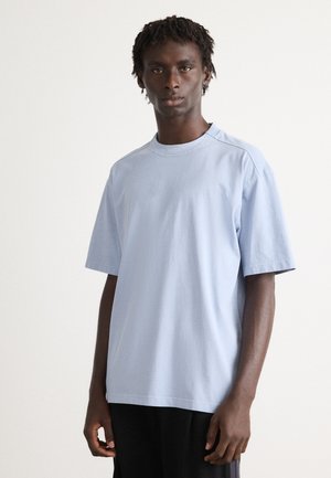 Young man with short dreadlocks wearing a light blue oversized T-shirt and black pants, standing against a plain white background.