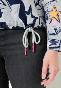 Grau melierter Pullover mit dunkelblauen Sternenprints, ausgestattet mit einem Kordelzugsaum, der mit hellgrauem Kordel und pinkfarbenen Kunststoffenden umwickelt ist, getragen über dunklen Jeans.