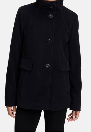 Femme portant un manteau noir boutonné avec poches latérales et col haut, debout devant un fond blanc uni.