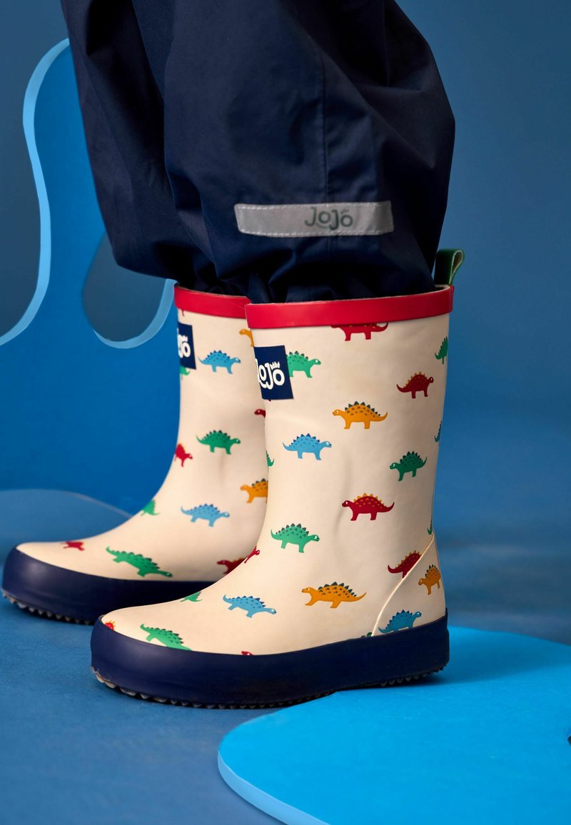 Enfant portant des bottes de pluie crème avec des imprimés colorés de dinosaures et une bordure rouge, debout sur un sol bleu devant un fond bleu.