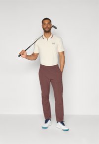 Bež majica s kratkim rukavima s burgundskim ovratnikom i logotipom, u kombinaciji s burgundskim hlačama. Model nosi bijele golf cipele s plavim akcentima i drži golf palicu.