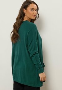 Maglione verde oversize con maniche lunghe, polsini a costine e scollatura rotonda. Tessuto morbido, design casual e schiena leggermente più lunga.