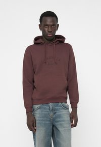 Maronfarvet hættejakke med en frontlomme, snørehætte og prægede logo. Sammen med lys blå slidte jeans. Glat stofstrukturen.