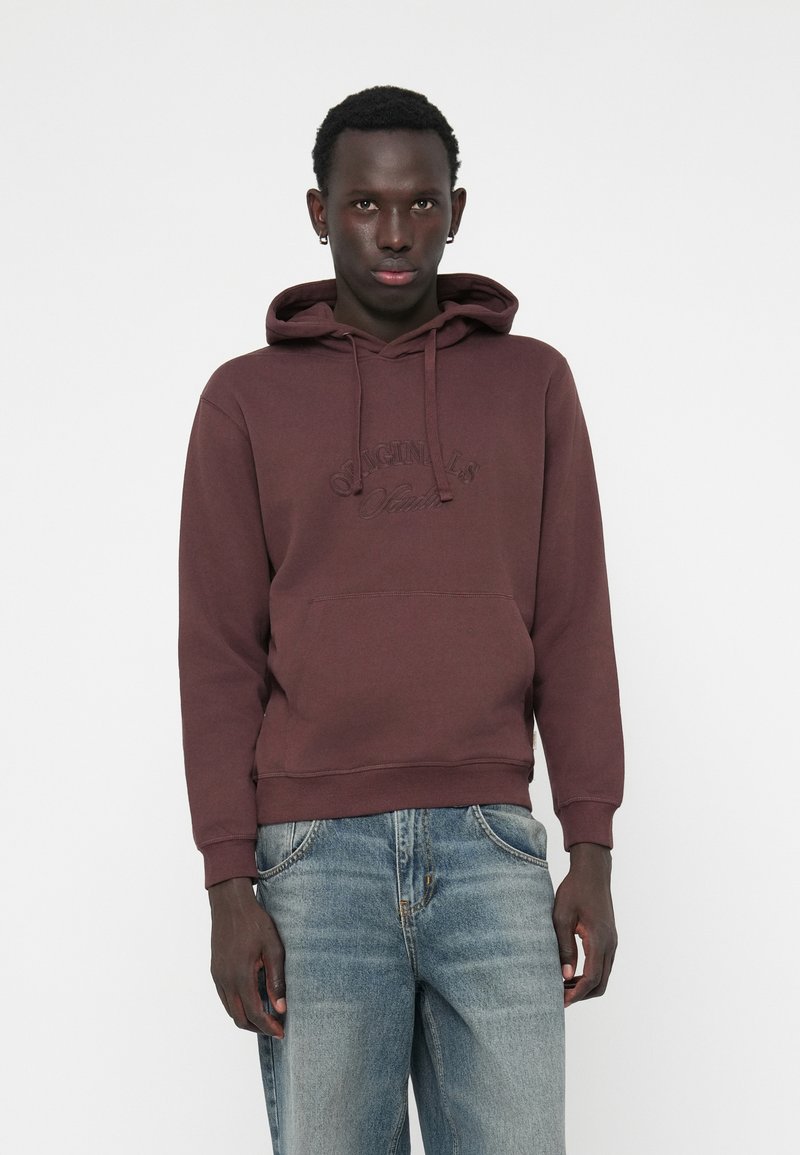 Maronfarvet hættejakke med en frontlomme, snørehætte og prægede logo. Sammen med lys blå slidte jeans. Glat stofstrukturen.