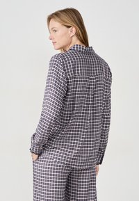 Koszula w kratę w granatowo-białym kolorze, z długimi rękawami, z zapięciem na guziki przy mankietach oraz detalem na plecach. Noszona z pasującymi spodniami w kratę.