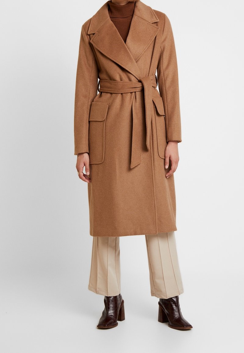 Femme portant un manteau camel ceinturé sur un pantalon beige et des bottines à talons brun foncé, sur un fond uni.