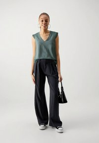 Zaļš v-veida kakla bezpiedurkņu tops un melnas augstās jostas bikses, kombinēts ar melnu somiņu un melnbaltām sporta kurpēm. Vienkāršs fons.