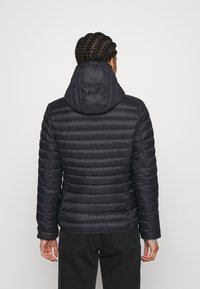 Person med fletninger iført en sort quilted jakke med hætte, stående med ryggen til mod en lys ensfarvet baggrund.