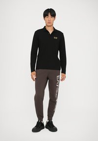 Juodas ilgomis rankovėmis polo marškinėliai su auksiniu logotipu, derinami su rudomis sportinėmis kelnėmis, ant kurių yra baltas užrašas. Modelis dėvi juodus sportinius batelius.