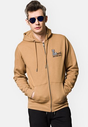 Beżowy hoodie na zamek z kapturem na sznurki, dwoma przednimi kieszeniami i haftowanym logo. Gładka tekstura, zestawiony z czarnymi spodniami i niebieskimi okularami przeciwsłonecznymi.