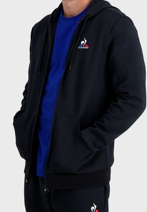 Mand iført sort lynlås-hoodie med Le Coq Sportif-logo over blå t-shirt, hænder i lommerne og sorte bukser med matchende logo.