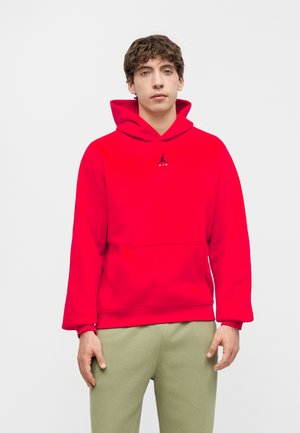 Jeune homme face à l’avant portant un sweat à capuche rouge vif avec un petit logo noir et un pantalon vert clair sur un fond uni.