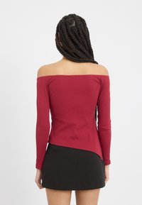 Burgunderfarget ribbestrikket avdelingsbluse med lange ermer og en asymmetrisk kant, kombinert med et svart skjørt. Teksturert stoff synlig.