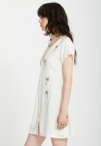 Robe blanche à manches courtes avec un décolleté en V, ornée d'un broderie de palmiers verts, avec un devant à boutons et une fente. Texture de tissu lisse.