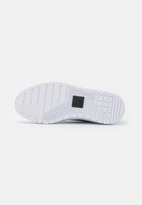 Semelle en caoutchouc texturé de chaussure blanche avec des rainures et un patch logo carré. Design épuré avec une couleur et un motif uniformes.
