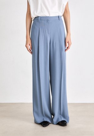 Personne portant un pantalon bleu clair taille haute à jambes larges avec passants de ceinture et des chaussures noires pointues, debout sur un sol clair contre un mur blanc.
