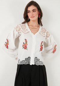 White cotton blouse with floral embroidery in red and green. Features lace inserts on sleeves and bottom hem, with a button front closure.