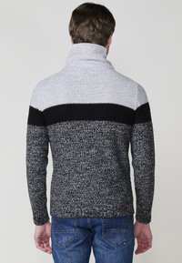 Grijze en zwarte gebreide trui met een hoge kraag, textuurstof en horizontale kleurblokken over de schouders en de rug.