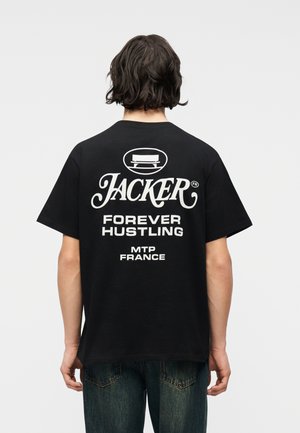 Oseba s srednje dolgimi temnimi lasmi, oblečena v črno majico s belim napisom "JACKER Forever Hustling MTP France" na hrbtu.