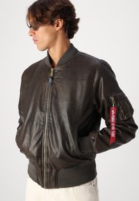 Jaqueta bomber de couro castanho com punhos e gola canelados, apresenta um fecho éclair na frente e um bolso lateral com uma etiqueta vermelha "REMOVER ANTES DO VOO".