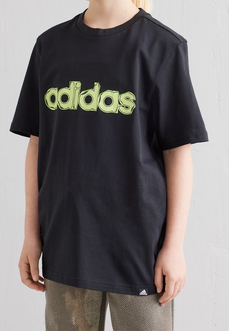Mustassa puuvillapaidassa on iso vihreä adidas-logo rinnassa. Lyhyet hihat ja pyöreä pääntie, yhdistettynä kuviollisiin housuihin.