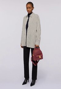 Pruhovaná oversized košile v béžové a bílé barvě, černé přiléhavé kalhoty a vínově červený batoh se zlatými kovovými doplňky. Černé lodičky na vysokém podpatku.
