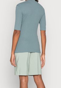 Hellblaues, geripptes Oberteil mit kurzen Ärmeln und hohem Ausschnitt, kombiniert mit hellgrünen, plissierten Shorts. Die Materialien wirken weich und bequem.