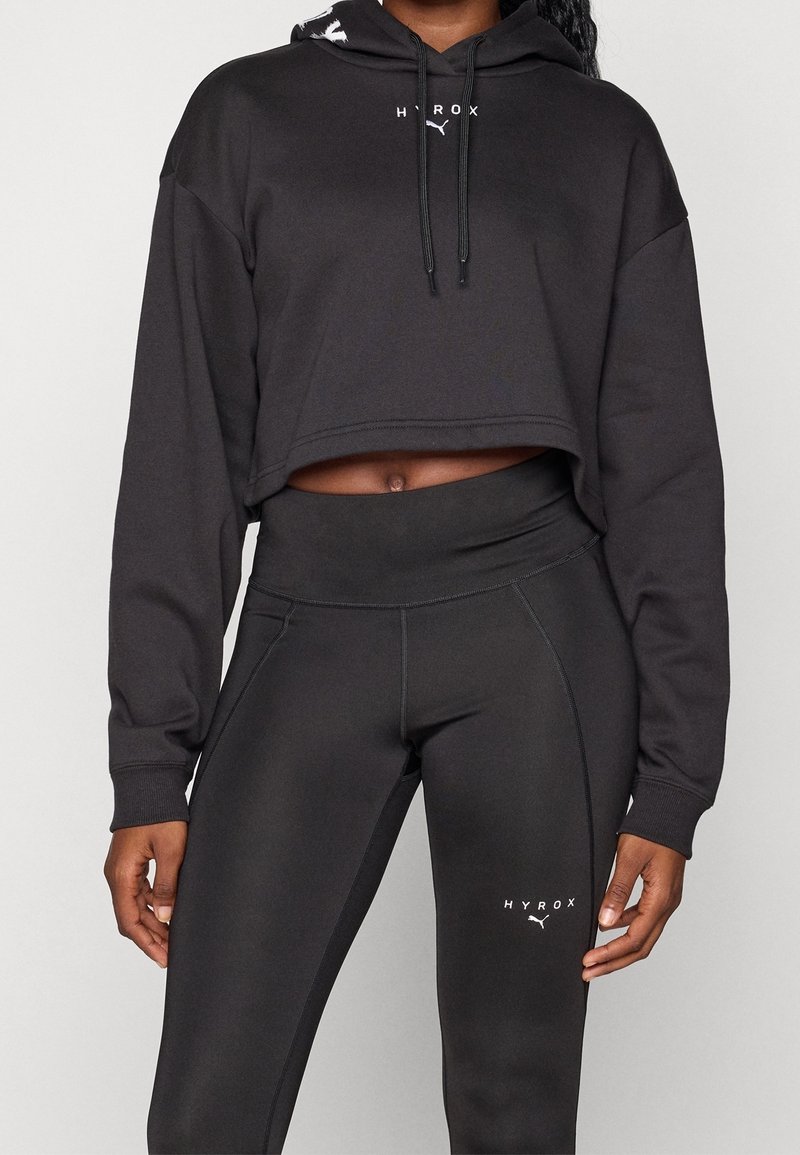 Personne portant un sweat à capuche court noir avec "HYROX" et logo Puma et des leggings noirs taille haute assortis avec "HYROX" et logo Puma.