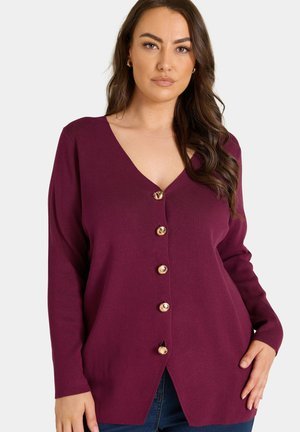Kvinde iført en langærmet lilla cardigan med guldknapper, stående med den ene hånd på hoften mod en ensfarvet baggrund.