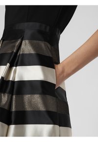 Gestreiftes schwarz-weißes Kleid mit abwechselnd matten und metallischen Stoffen. Verfügt über eine Tasche und eine glatte, figurbetonte Silhouette.