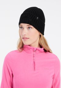Berretto a maglia nero con motivo a coste verticali, caratterizzato da un accento del logo. Indossato con un top in pile rosa a zip quarti, che evidenzia il contrasto di texture.