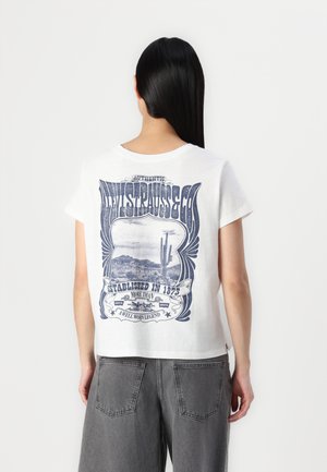 T-shirt en coton blanc avec un design graphique bleu comportant du texte et un paysage. Manches courtes, col rond, coupe décontractée. Un pantalon gris est visible.