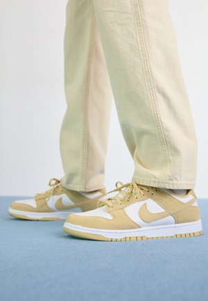 Person iført beige og hvide Nike-sneakers med matchende beige bukser, stående på en lyseblå gulv.