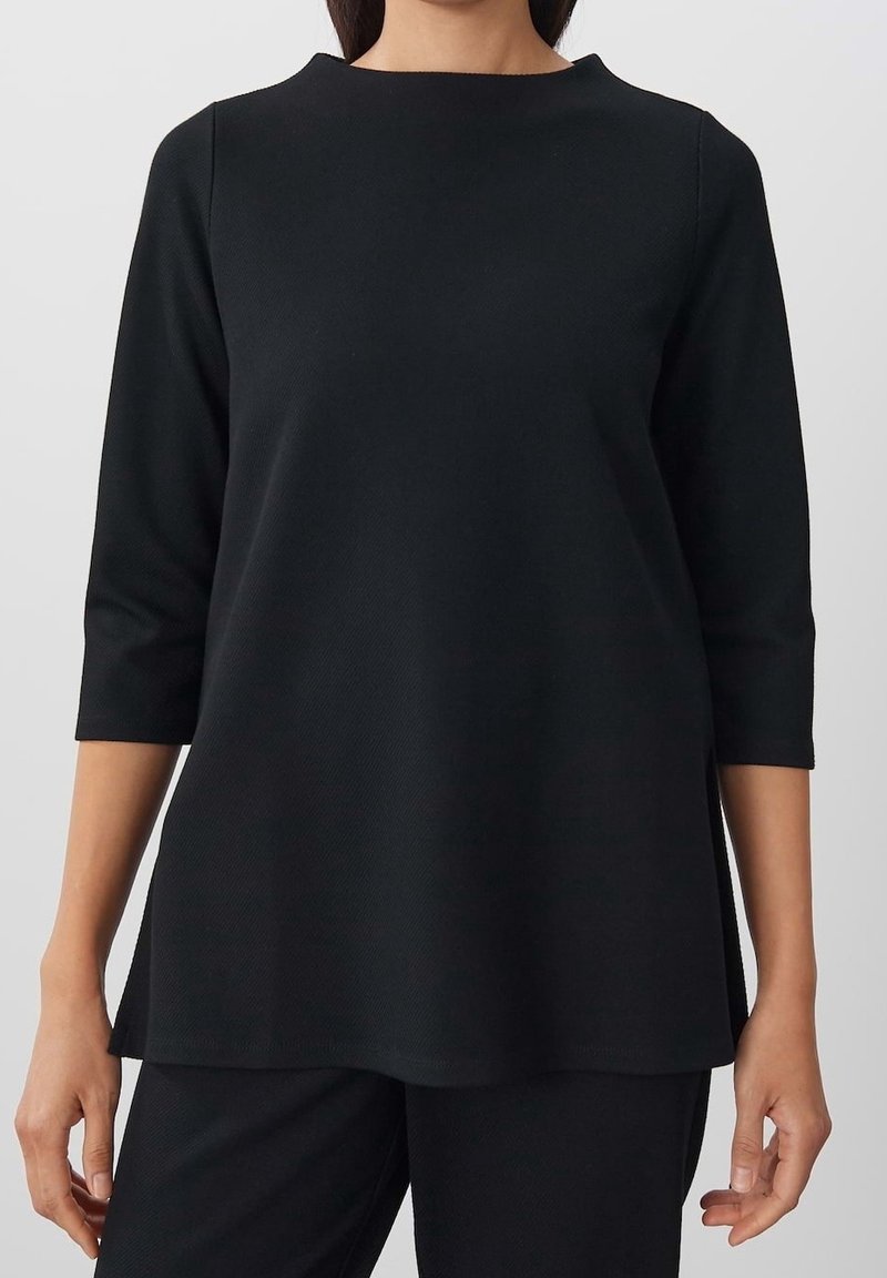 Femme portant un haut noir ample à manches trois quarts et un pantalon noir assorti sur un fond uni.