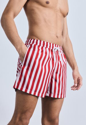 Άνδρας χωρίς πουκάμισο, φορώντας κόκκινα και λευκά κατακόρυφα ριγέ μαγιό με ελαστική μέση και κορδόνι, με το ένα χέρι στην τσέπη.